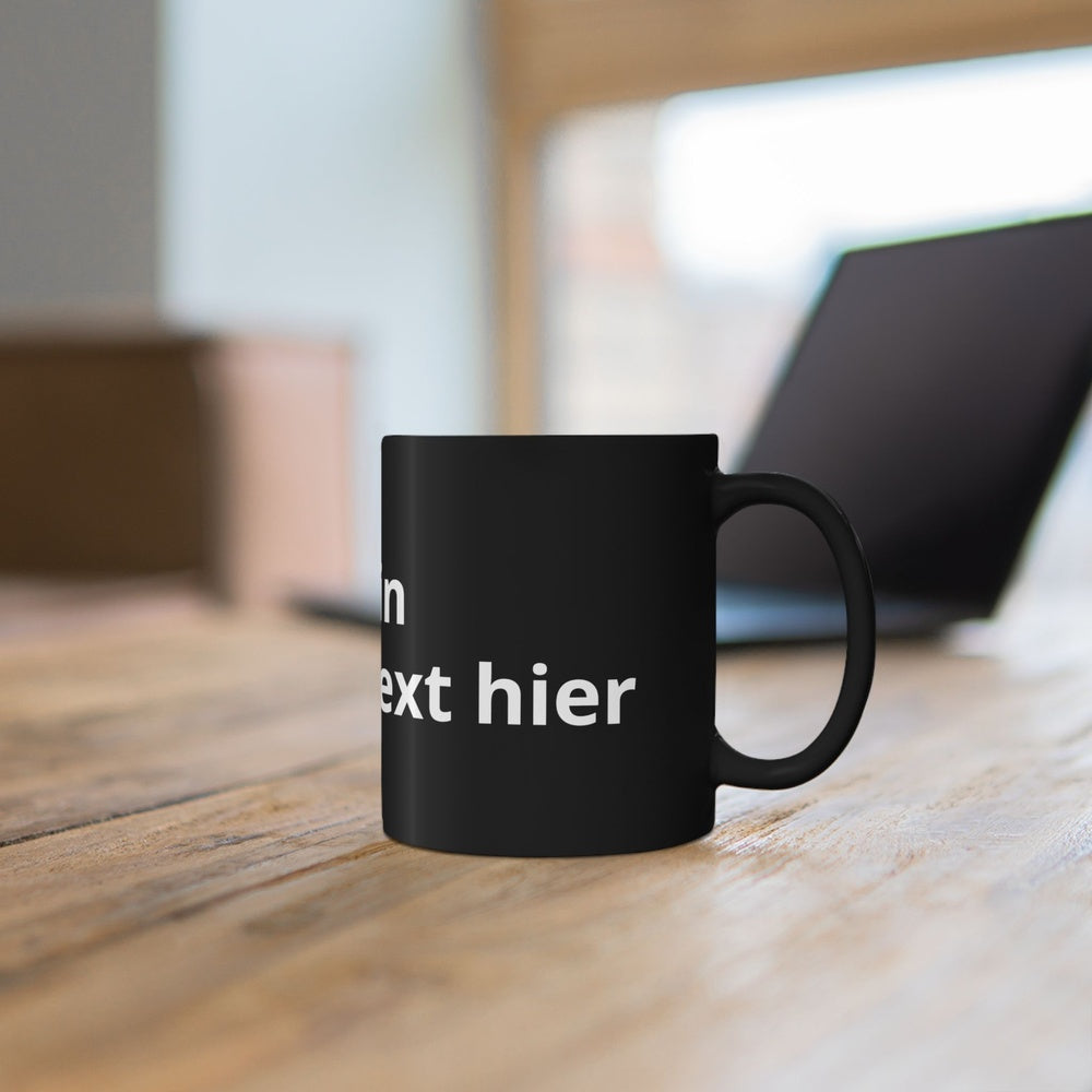 Schwarze Keramiktasse mit individuellem Wunschtext auf einem Holztisch vor einem Laptop.